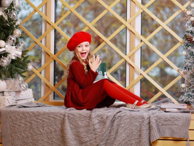 Kırmızılı Sarışın Kız, Pencere kenarındaki Elinde Bir Hediyeyle, Bir Noel ağacının yanındaki camın ötesine bakıyor