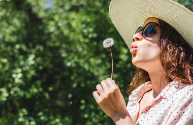 Şapka takan ve güneş gözlüğü takan genç mutlu kıvırcık kadın doğada bir karahindibaya üflüyor..