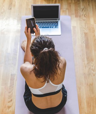 Yoga minderinde oturan ve cep telefonunu kullanan bir kadın..