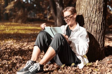 Dişlerinde diş teli olan, kitap okuyan ve parkta otururken gülümseyen genç, kısa saçlı kadın..