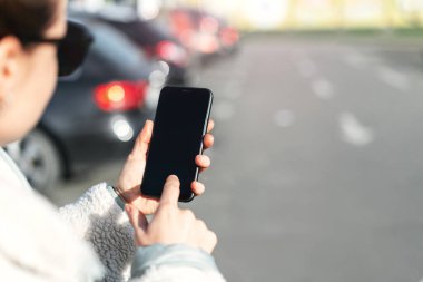Akıllı telefonunu kullanan bir kadın, otoparkta dikilirken parmaklarıyla ekrana dokunuyor..