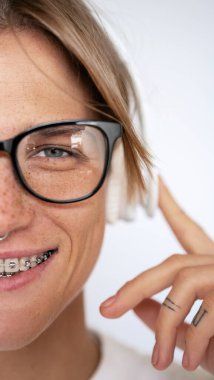 Cropped portrait woman in dental brakes glasses listening music with headphones looking at camera half face against white background. 