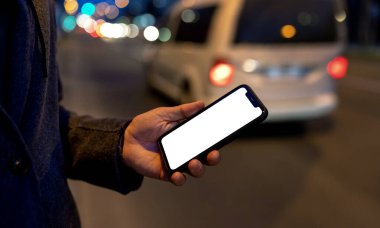 Smartphone with blank screen in male hand in city at night, ride hailing service mobile app.