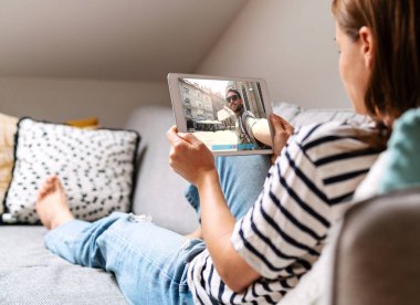 Adam seyahat ediyor ve kız arkadaşını videoya çekiyor. Evdeki kanepede uzanmış dijital tablet kullanarak sevgilisiyle sohbet eden bir kadın..