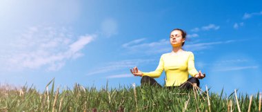 Mavi gökyüzüne karşı yeşil çimlerde otururken dışarıda yoga egzersizi yapan genç bir kadın..