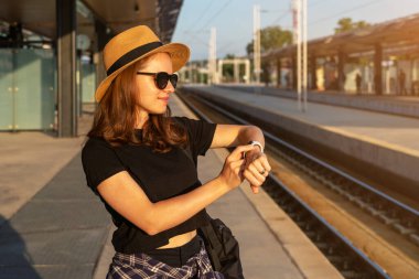 Hasır şapka takan turist kadın tren istasyonunda bekliyor ve akıllı saatini kullanıyor..