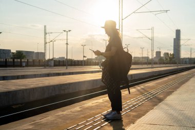 Sırt çantalı turist kadın tren istasyonunda duruyor ve gün batımında telefon kullanıyor..