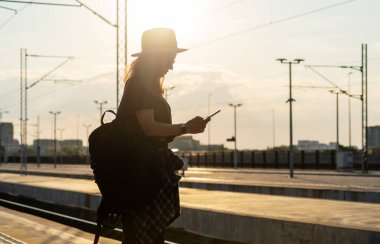 Sırt çantalı turist kadın tren istasyonunda duruyor ve gün batımına karşı telefonu elinde tutuyor..