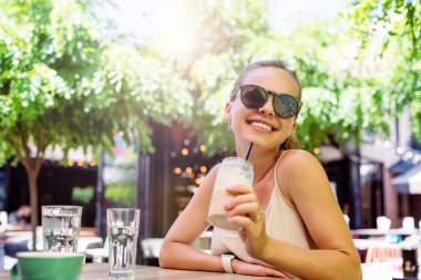 Güneş gözlüklü mutlu esmer kadın sokak kafesindeki masada oturuyor ve buzlu kahve içiyor..