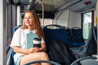 Kamu otobüsünde oturan genç kız öğrenci elinde termal bardak ve çalışma kitaplarıyla pencereden dışarı bakıyor..