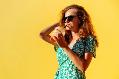 Sexy kendinden emin, yeşil elbiseli ve güneş gözlüklü kadın yazın sarı duvarın yanında poz veriyor, samanla içki içiyor ve gülümsüyor..