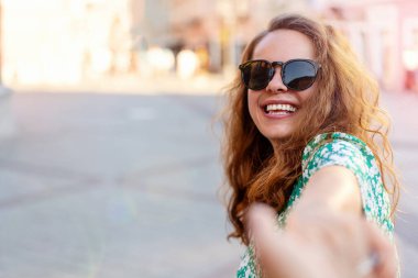 Genç, kıvırcık bir kadın partnerinin elini çeker, kameraya bakar ve güler..