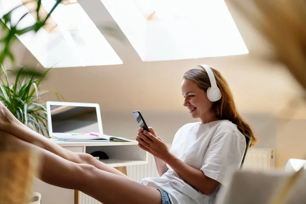 Young woman wearing white clothes and white headphones sits with her feet up on the table uses her smart phone and smiling.