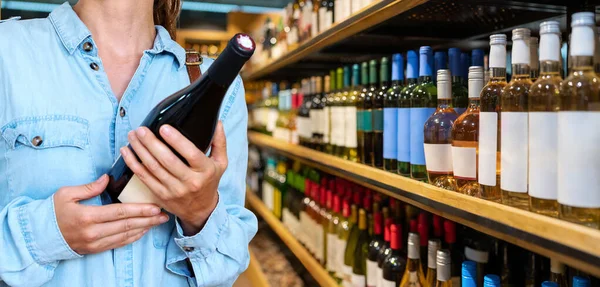Female customer holding bottle of red wine in hands on background of shelves of wine in store.