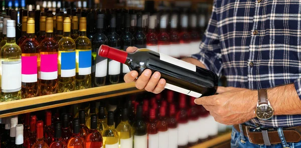 Customer in shop holding bottle of wine in his hands and reading information about the wine on the label.