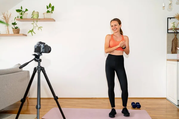 Young athletic woman fitness coach wearing sportswear standing in front of camera on tripod and recording her video blog about healthy lifestyle.