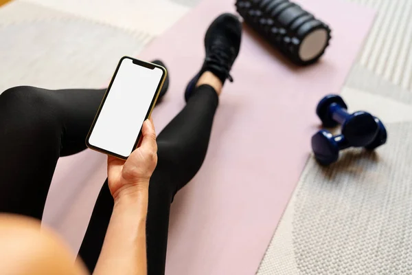Female sports person sits on yoga mat and holding smart phone with white empty screen in her hand. Mobile phone and fitness. Workout mobile app.