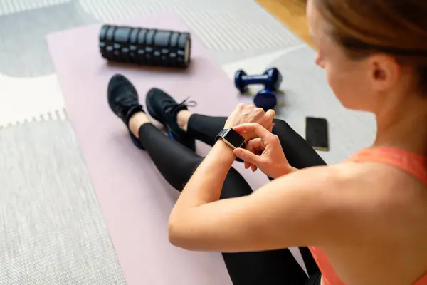 Athletic woman wearing sportswear woman sits on exercise mat and launching training program on her smart watch. Fitness and personal digital devices..