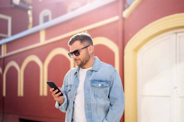 Güneş gözlüklü ve kot ceketli yakışıklı genç adam sokakta yürürken cep telefonuna bakıyor..