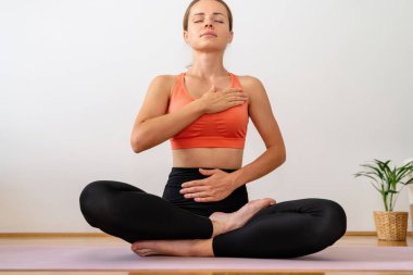 Genç yetişkin bir kadın yoga minderinde lotus pozisyonunda oturuyor ve nefes egzersizi yapıyor..