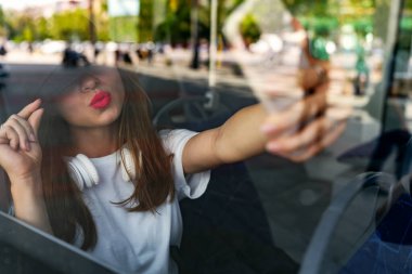 Şehir otobüsünde otururken selfie çeken şık şehirli kadın. Şehir hayatı, camdan fotoğrafçılık. Cep telefonu mesajlaşması.