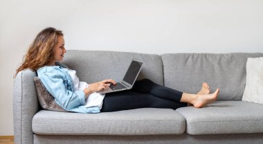 Genç esmer kadın, gündelik giysiler giyip evdeki kanepede uzanıyor ve laptopunu kullanıyor. İnternetten alışveriş, elektronik bankacılık, e-ticaret..