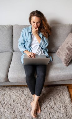 Genç esmer kadının yüksek açılı görüntüsü evdeki koltukta oturuyor ve laptopunu kullanıyor..