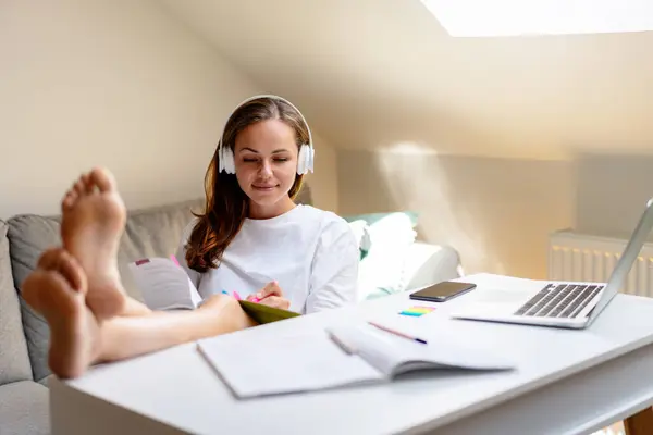 Self-development. Attractive brunette woman in headphones using workbook at home desk. Self-directed learning.