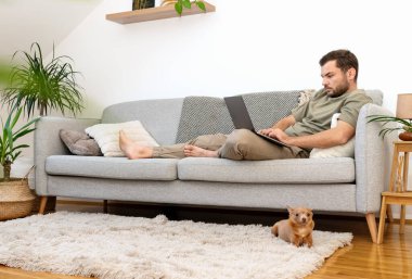 Evdeki kanepede yatıp dizüstü bilgisayarını kullanan bir adam. Evden çalışıyorum. Yurt içi yaşam tarzı.