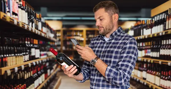 Πελάτης Επιλέγει Κρασί Στην Κάβα Χρησιμοποιώντας Smartphone Του Για Σάρωση Εικόνα Αρχείου