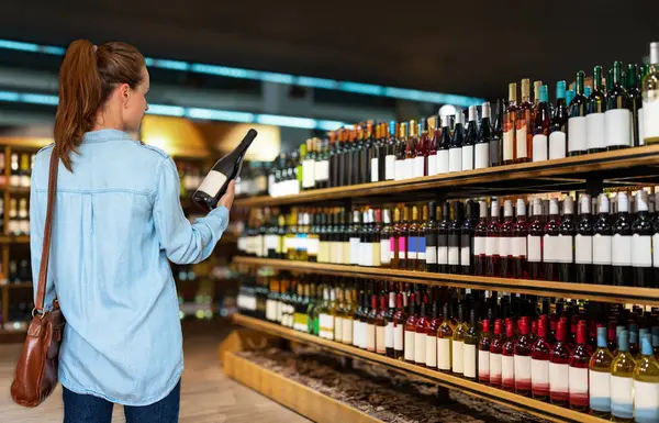 Rear view of woman customer stands in front of shelves with wine bottles choosing wine in liquor store.