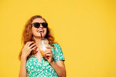 Yaz yeşili gözlüklü, meyve suyu içen ve sarı duvarın önünde poz veren mutlu genç kadın. Kadınlar yaz günlerinde eğleniyor..