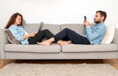 Genç bir çift evdeki kanepede dinleniyor ve kişisel dijital cihazlarını kullanıyorlar..