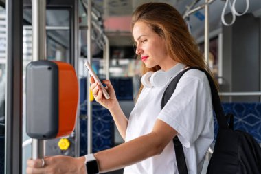 Beyaz tişörtlü esmer kadın otobüsün içinde duruyor ve akıllı telefonunu kullanıyor..