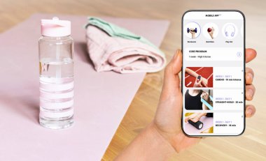 Mobil spor uygulaması ile ev antrenmanı. Yoga minderinin, havlunun ve su şişesinin önündeki ekranda spor mobil uygulama arayüzü.