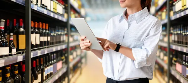 Woman retail sales manager analysing commercial performance of alcohol department in supermarket. Female employee using digital tablet in liquor store.