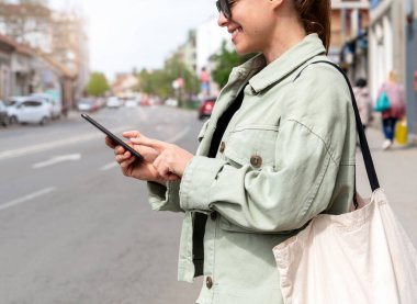 Kadın, akıllı telefonunu kullanarak sokakta araba paylaşımı ya da araba kiralama aracı arıyor. Kadın şehirde yol kenarında duruyor ve telefonunu taksi aramak ya da beklemek için kullanıyor..