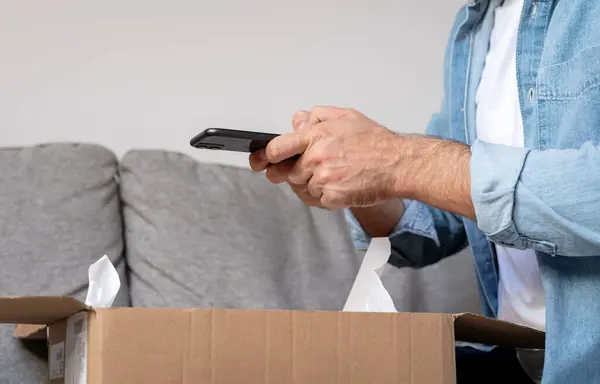 Male person unpacking parcel cardboard and takes a photo of his purchase, internet order. Person sharing feedback with online store using his smartphone.