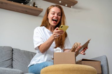 Neşeli kadın internetten alışveriş yapıyor ve kartondaki malların fotoğrafını çekiyor. Evden alışveriş ve hızlı teslimat..