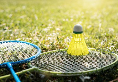 İki badminton raketi ve yeşil çimlerin üzerinde sarı mekikli horoz. Yaz tatili ve spor aktiviteleri.