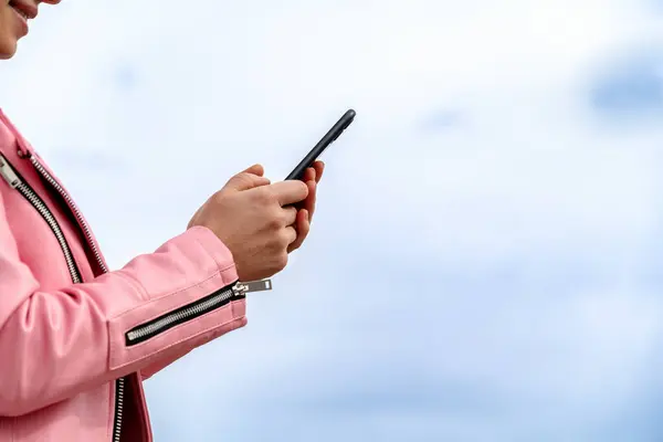 Smartphone in woman\'s hands in front of sky. Female in pink jacket messaging online wile waking outdoors.