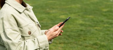 Bir kadın açık havada yeşil bir çimenliğin arka planına karşı cep telefonu kullanıyor. Kadının elindeki akıllı telefonun yakın plan fotoğrafı..