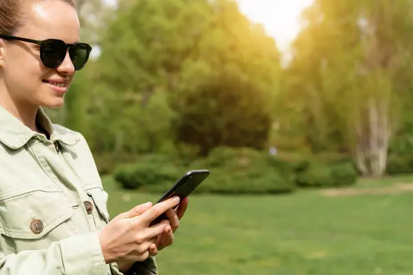Woman in sunglasses messaging online using smartphone while walking outdoors in a city park.