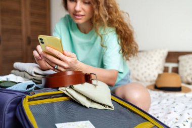 Uçak bileti alan ve otelde rezervasyon yapan bir kadın cep telefonunu evde kullanıyor ve tatil için seyahat çantası hazırlıyor..