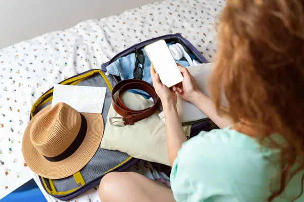 Woman packing her travel bag and checking her checklist on her smartphone. Preparation for traveling, booking or reservation hotel and flight tickets.