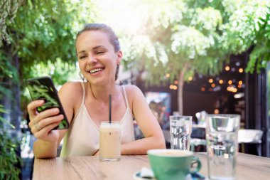 Esmer kadın yazın bir kafede dinleniyor ve internette akıllı telefon kullanarak sohbet ediyor..
