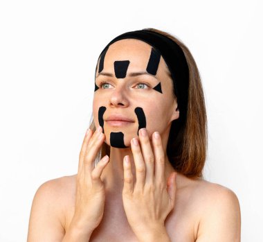 Female beauty model applying kinesio facial tapes on her face against a white background in studio. Facial contouring and skin tightening.
