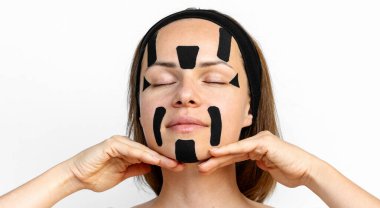 Woman beauty model applying black coloured kinesio facial tapes on her face on white background. Facial contouring and skin tightening.