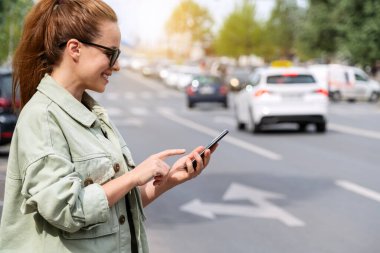 Şehir kadını şehir yolunun yanında dikilirken akıllı telefonunu kullanıyor. Taksi çevrimiçi servisi. Hizmet olarak hareket kabiliyeti. Araba paylaşım kavramı.