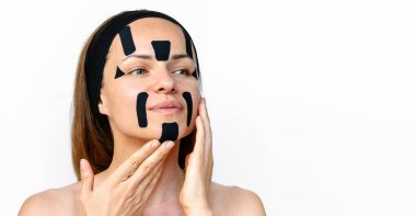 Female beauty model applying kinesio facial tapes on her face against a white background in studio.. Facial contouring and skin tightening.